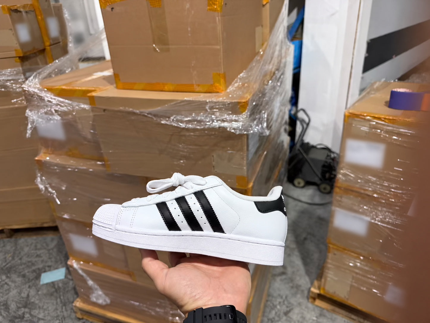 A hand holding an Adidas sneaker in front of stacked shipping cartons in China